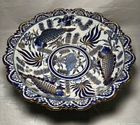 Top view of a large round porcelain pedestal bowl with scalloped edges featuring hand painted blue fish and floral patterns accented with gold.