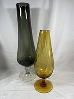 Pair of two mid century modern hand blown glass pedestal vases, one yellow/amber and one smokey gray, on a white backdrop.