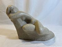 Side view of white resin sculpture of reclining nude woman with gray speckles, showing curved form and bent arms raised behind head.