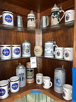 Wide view of corner shelf showing multiple beer steins, NASA logo mugs, and mugs with crests arranged grouped by type.