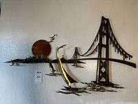Full view of metal wall sculpture showing bridge span, skyline, copper sun disk, waves and birds