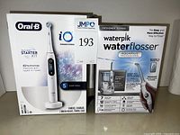 Boxed Oral-B iO rechargeable toothbrush starter kit and Waterpik Water Flosser boxes side by side.