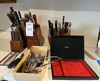 Photo of three wooden knife blocks holding multiple knives, a tray with assorted knives and kitchen shears, and an empty steak knife case opened, showing red velvet interior.