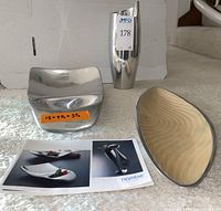 Three Nambe items shown together on carpet. Includes tall polished vase, square metal wave shaped dish, and oblong cream dish with metal rim. Included Nambe brochure also visible.