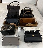 Photo showing nine vintage handbags arranged in a box, different styles, mostly black with one brown and one white bag.