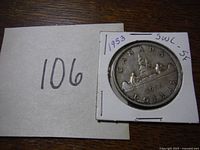 Photo of the reverse side of the 1953 Canadian silver dollar coin in cardboard holder with handwritten notes and numerical tag 106 card next to it.