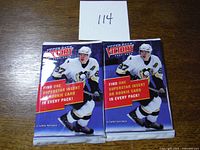 Front view of two unopened 2007-08 Upper Deck Victory hockey card packs on a wooden surface with a note labeled '114'. The packs display a hockey player in white and black gear and the text about finding a superstar insert or rookie card in every pack.
