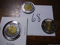 Three Canadian twoonie coins displayed on a wooden surface—two loose and one in a protective holder, featuring war hero designs
