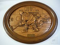 Full view of the oval wooden plaque carved with a hockey scene of Wayne Gretzky and a goaltender on the ice near a net with arena advertisements and a banner.