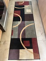 Runner rug with geometric abstract design in muted colors of black, gray, beige, brown, purple, burgundy, and red, featuring curved lines and block shapes