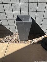 Photo showing one tall, square, dark gray planter outdoors against a gray concrete block wall on a stone surface.