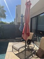 Two outdoor patio umbrellas in closed position with metal frames, one coral red and one slate blue, placed near patio chairs and fence.