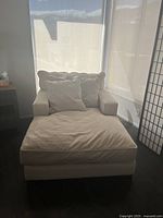 Front view of the beige upholstered lounge chair with two matching cushions, placed near window with white shades.