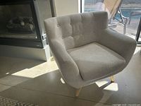 Full view of a light beige or grey fabric upholstered armchair with button tufted backrest and wooden legs. The chair is positioned near a glass door with a pool visible outside.