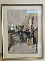 Framed abstract painting with neutral and yellow tones, displayed on a wall with light reflecting on the glass.