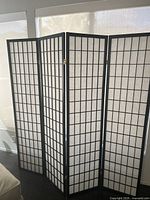 Four-panel black framed folding screen with white translucent inserts arranged in a grid pattern standing upright near a window