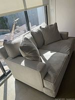 Front and side view of grey fabric sofa with three large back cushions and two seat cushions positioned near a large window letting in natural light.