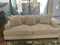 Front view of beige fabric sofa with four loose back cushions and two armrest cushions placed in room with glass door and patio outside