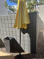 Yellow outdoor patio umbrella closed, shown against a stone wall, standing in a base container surrounded by small decorative rocks, under natural daylight.