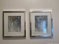 Two framed abstract paintings side by side on a wall showing textured mixed color palette and silver frames