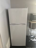 Front view of white Haier refrigerator with freezer top and fridge bottom resting against wall. Adjacent metal shelf and microwave visible.
