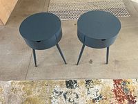 Photo showing two modern round side tables in a dark gray/blue color with three legs each and a single drawer, placed on a floor with patterned rugs nearby.