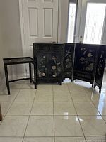 Full shot of the set including the black console cabinet with floral paintings and brass hardware, the black side table, and the three-panel black folding screen with floral designs, all arranged on tiled floor near front door.