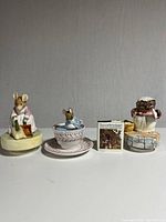 Group photo of three ceramic music boxes and small Peter Rabbit book together on a white surface.