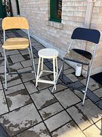 Three stools pictured outside on tiled ground near brick wall. Two are collapsible high stools, one with natural wood seat and backrest, the other with black seat and backrest; the third is a small round white seat stool.
