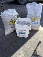 Two large bags and one bucket containing fine gravel placed on pavement with a truck in the background.