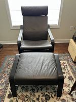 Front view of black leather armchair with matching stool placed on patterned rug near window, showing overall condition and design.