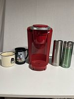 Red Keurig coffee maker centered with two ceramic coffee mugs on the left and two Contigo travel mugs on the right on a white surface against white background.