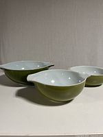 Front view of three Pyrex mixing bowls of different sizes arranged on flat surface, showing their green exterior and white interior with pouring spouts and handles visible.