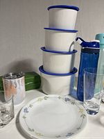 Four white stacking Tupperware bowls with blue lids in descending size order, surrounded by plates, glasses, and water bottles on a white surface