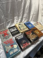 Eight hardcover vintage books laid on a white fabric background showing dust jackets: Camp X, Inside Camp X, Shutting Down the National Dream, War and Remembrance by Herman Wouk, The Winds of War by Herman Wouk, Hotel by Arthur Hailey, The Three Pillars of Zen, Dawn in the West, and Men Are from Mars Women Are from Venus.