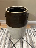 Photo of large two-tone crock with handles on a carpeted floor against beige wall.