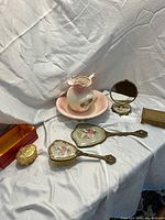 Full view of dresser set on white draped fabric background showing hand mirror, brush, pink pitcher and basin, small round compact mirror, rectangular gold-tone box and a gold decorative item in red box.