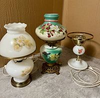 Three vintage hurricane lamps displayed side by side on floor against a wooden wall.