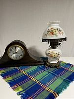Photo of the vintage wooden mantel clock next to a white glass floral hurricane lamp on a blue and green plaid cloth