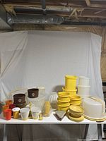 Full view of all vintage Tupperware items displayed on a white table, showing various containers, bowls, cups, and kitchenware in multiple colors including yellow, brown, cream, orange, and white.