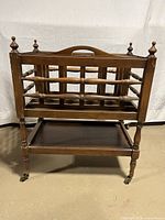 Front view of the wooden vintage magazine rack showing spindle-style rails and finials on corners, lower shelf visible, on wheels.