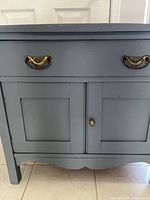 Front view of the wash stand showing light blue painted wooden surface, drawer with brass handles, and cabinet doors with round brass knobs.