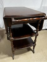 Front angled view showing side drop leaf folded down, two shelves, turned legs, and front caster wheels.