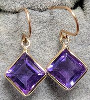 Close-up natural light photo of 10K yellow gold earrings with square amethyst stones placed on gray fabric.