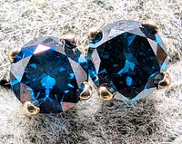 Close-up of the pair of round cut lab-created blue diamond earrings with 14K white gold prong settings showing deep blue color and clarity.