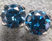 Close-up of one earring showing round brilliant cut blue diamond in four-prong white gold setting