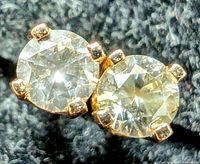 Close-up photo of one yellow gold stud earring with a single round brilliant cut diamond held by four prongs on a dark fabric background.