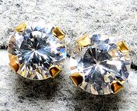 Close-up of a pair of yellow gold stud earrings with round clear 4mm cubic zirconia stones secured by 4 prongs, displayed on grey fabric.