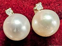 Close-up front view of the pair of 14K yellow gold earrings with 5.5mm round freshwater pearls and small round diamonds on top against a red fabric background.