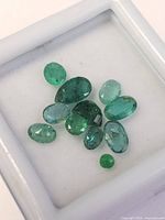 Close-up of 11 loose emerald gemstones in a white display box on a white background, showing a variety of shapes and green shades.